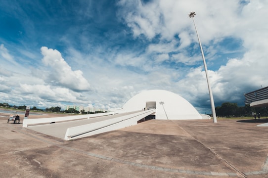 A large, modern white dome building with a long ramp leading up to its entrance is situated in an open area. The sky is partly cloudy with patches of blue showing through. Surrounding the building, there are some trees and distant buildings, along with a tall light pole. The ground appears paved and the scene has an overall expansive feeling.