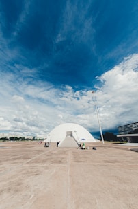 A white, dome-shaped building with a ramp entrance labeled 'MUSEU' is centered in an open, expansive concrete area. People are scattered around, some walking, some seated under a blue umbrella. The sky above is vast and dramatic, filled with a mix of blue and white clouds. A tall lamppost stands to the right, and modern buildings are visible in the background.