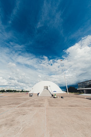 A white, dome-shaped building with a ramp entrance labeled 'MUSEU' is centered in an open, expansive concrete area. People are scattered around, some walking, some seated under a blue umbrella. The sky above is vast and dramatic, filled with a mix of blue and white clouds. A tall lamppost stands to the right, and modern buildings are visible in the background.