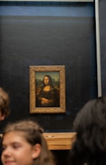 A famous painting is displayed in a gallery setting, framed in a decorative gold frame. The background is dark, allowing the painting to stand out prominently. In the foreground, there are blurred individuals likely observing the artwork.