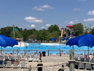 A bustling water park scene with numerous people swimming in a large wave pool. Several water slides, including a prominent red and yellow one, are visible in the background. Bright blue umbrellas and rows of empty lounge chairs are scattered around the pool deck. Lush green trees surround the area under a partly cloudy sky.