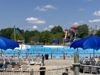A bustling water park scene with numerous people swimming in a large wave pool. Several water slides, including a prominent red and yellow one, are visible in the background. Bright blue umbrellas and rows of empty lounge chairs are scattered around the pool deck. Lush green trees surround the area under a partly cloudy sky.