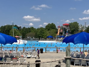 A bustling water park scene with numerous people swimming in a large wave pool. Several water slides, including a prominent red and yellow one, are visible in the background. Bright blue umbrellas and rows of empty lounge chairs are scattered around the pool deck. Lush green trees surround the area under a partly cloudy sky.