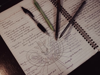 A spiral-bound notebook with handwritten notes and a detailed anatomical drawing. Three pens with different colored inks are placed across the open pages, each with studied outlines and annotations.