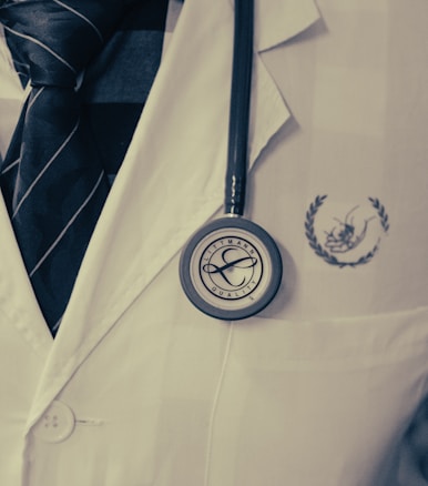 A close-up of a person wearing a white coat and a dark tie, with a stethoscope hanging from the neck. The stethoscope is positioned over the chest area, displaying the brand name 'Littmann' clearly. The coat has a logo on it.