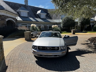 A silver convertible car is parked in a circular driveway in front of a large, stone house with a complex roof structure and several dormer windows. The area is landscaped with trees and ornamental grasses, and the driveway is made of interlocking pavers.