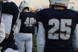 A group of American football players wearing navy jerseys with white numbers stand on a field. Helmets with white and dark blue details are worn by the players, and their backs are turned to the camera.