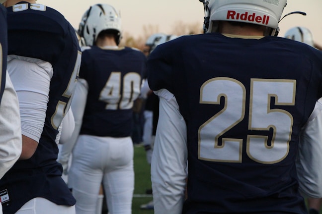 A group of American football players wearing navy jerseys with white numbers stand on a field. Helmets with white and dark blue details are worn by the players, and their backs are turned to the camera.