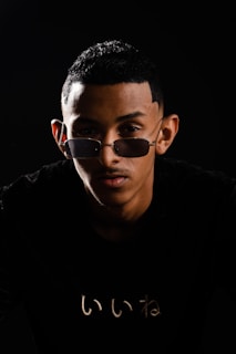A person with short black hair and sunglasses is positioned against a dark background. The lighting creates dramatic shadows, emphasizing facial features and the reflective nature of the glasses. A subtle graphic or text is visible on the black shirt worn by the person.
