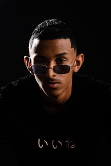 A person with short black hair and sunglasses is positioned against a dark background. The lighting creates dramatic shadows, emphasizing facial features and the reflective nature of the glasses. A subtle graphic or text is visible on the black shirt worn by the person.