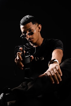 A person wearing sunglasses and a black shirt is holding a walkie-talkie. The image has a dark and dramatic atmosphere, with the subject sitting and extending the device toward the camera.