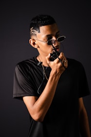 A person wearing sunglasses with unique hair design holds a two-way radio close to their mouth, dressed in a black t-shirt and sporting a gold chain. The background is a dark, neutral color that emphasizes the figure.
