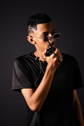 A person wearing sunglasses with unique hair design holds a two-way radio close to their mouth, dressed in a black t-shirt and sporting a gold chain. The background is a dark, neutral color that emphasizes the figure.