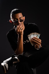 A person wearing sunglasses sits on a chair holding a wad of cash in one hand while talking on a phone held to the ear with the other hand. They are dressed in casual black clothing and sneakers, set against a dark background.
