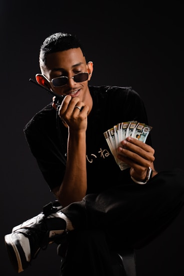 A person wearing sunglasses sits on a chair holding a wad of cash in one hand while talking on a phone held to the ear with the other hand. They are dressed in casual black clothing and sneakers, set against a dark background.