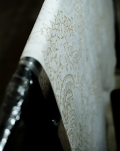 A close-up view of a roll of wallpaper with intricate floral patterns. The design is subtle, featuring soft, light outlines against a cream background. The wallpaper is partially unrolled and draped across a surface, with the focus on the details of the pattern. The lighting casts gentle shadows, emphasizing the texture.