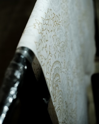 A close-up view of a roll of wallpaper with intricate floral patterns. The design is subtle, featuring soft, light outlines against a cream background. The wallpaper is partially unrolled and draped across a surface, with the focus on the details of the pattern. The lighting casts gentle shadows, emphasizing the texture.