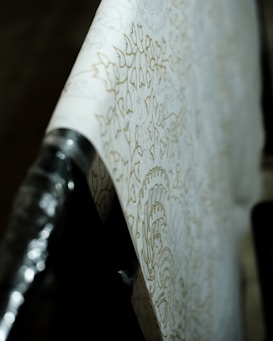 A close-up view of a roll of wallpaper with intricate floral patterns. The design is subtle, featuring soft, light outlines against a cream background. The wallpaper is partially unrolled and draped across a surface, with the focus on the details of the pattern. The lighting casts gentle shadows, emphasizing the texture.