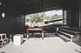 A covered patio area features a wooden table with two chairs, a cooler, and some small objects on the surface. The space includes steps leading up to a door, and is surrounded by dark walls. String lights hang from the ceiling, and a circular mirror is mounted on the wall. In the background, trees and part of a house roof are visible.