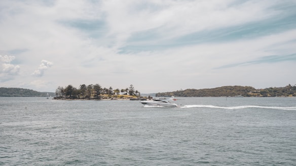 A motorboat cruises along calm waters with a trail of white wake behind it. In the distance, a small island covered in trees is visible under a cloud-streaked sky, creating a serene maritime landscape.