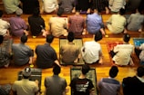 A group of people are sitting on prayer mats in a place of worship. They are positioned closely together in multiple rows facing the same direction. The setting suggests that they are engaged in a communal prayer session.