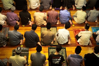 A group of people are sitting on prayer mats in a place of worship. They are positioned closely together in multiple rows facing the same direction. The setting suggests that they are engaged in a communal prayer session.