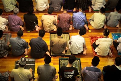 A group of people are sitting on prayer mats in a place of worship. They are positioned closely together in multiple rows facing the same direction. The setting suggests that they are engaged in a communal prayer session.