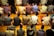 A group of people are sitting on prayer mats in a place of worship. They are positioned closely together in multiple rows facing the same direction. The setting suggests that they are engaged in a communal prayer session.