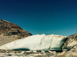 A large glacier rests amidst barren rocky mountains under a clear blue sky. The glacier appears to be slightly melting, with patches of ice on the ground in front of it.