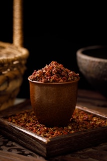 A rustic brown cup filled with red Sichuan peppercorns is placed on a wooden tray, which is also scattered with more peppercorns. In the background, a wicker basket and a stone bowl are partially visible, giving the scene a warm and earthy ambiance.