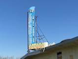 A large vertical sign with blue background lists a variety of items including 'Farm Supplies,' 'Farm Tools,' 'Hardware,' and 'Feed.' The sign features a retro design with a smaller section at the bottom indicating 'Honey Mist Beauty' surrounded by a honeycomb pattern. It is mounted on the roof of a building under a clear blue sky.