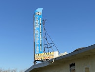 A large vertical sign with blue background lists a variety of items including 'Farm Supplies,' 'Farm Tools,' 'Hardware,' and 'Feed.' The sign features a retro design with a smaller section at the bottom indicating 'Honey Mist Beauty' surrounded by a honeycomb pattern. It is mounted on the roof of a building under a clear blue sky.