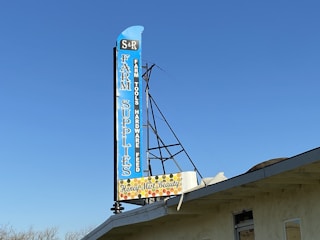 A large vertical sign with blue background lists a variety of items including 'Farm Supplies,' 'Farm Tools,' 'Hardware,' and 'Feed.' The sign features a retro design with a smaller section at the bottom indicating 'Honey Mist Beauty' surrounded by a honeycomb pattern. It is mounted on the roof of a building under a clear blue sky.