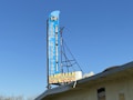 A large vertical sign with blue background lists a variety of items including 'Farm Supplies,' 'Farm Tools,' 'Hardware,' and 'Feed.' The sign features a retro design with a smaller section at the bottom indicating 'Honey Mist Beauty' surrounded by a honeycomb pattern. It is mounted on the roof of a building under a clear blue sky.