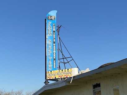 A large vertical sign with blue background lists a variety of items including 'Farm Supplies,' 'Farm Tools,' 'Hardware,' and 'Feed.' The sign features a retro design with a smaller section at the bottom indicating 'Honey Mist Beauty' surrounded by a honeycomb pattern. It is mounted on the roof of a building under a clear blue sky.