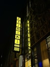 A tall building displays a vibrant, illuminated sign with the word 'FABRIKA' and 'HOSTEL' vertically aligned. The sign is bright yellow, set against a dark night background. The surrounding structure features large windows and a detailed decorative facade.