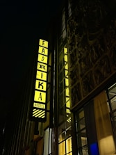 A tall building displays a vibrant, illuminated sign with the word 'FABRIKA' and 'HOSTEL' vertically aligned. The sign is bright yellow, set against a dark night background. The surrounding structure features large windows and a detailed decorative facade.