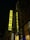 A tall building displays a vibrant, illuminated sign with the word 'FABRIKA' and 'HOSTEL' vertically aligned. The sign is bright yellow, set against a dark night background. The surrounding structure features large windows and a detailed decorative facade.