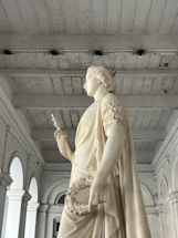 A white marble statue of a female figure adorned with a crown, holding a scepter and a laurel wreath. The setting is a grand architectural space with high ceilings and arched windows. The statue exudes a sense of grace and authority.