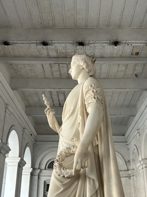 A white marble statue of a female figure adorned with a crown, holding a scepter and a laurel wreath. The setting is a grand architectural space with high ceilings and arched windows. The statue exudes a sense of grace and authority.