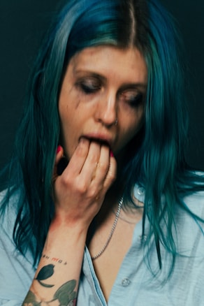 A person with long teal hair appears to be in a distressed or emotional state. Wearing a light blue shirt, they have smudged makeup around their eyes. A tattoo with floral design is visible on their arm as they place their fingers in their mouth.