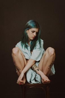 A woman with blue hair sits on a wooden chair, wrapping herself in a light blue towel. She appears contemplative, with her arms wrapped around her knees and tattoos visible on her arms and legs. The background is dark and minimalistic.
