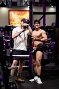 Two individuals in a gym setting, one is taking a photo, and the other is posing with a smile, wearing athletic gear.