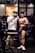 Two individuals in a gym setting, one is taking a photo, and the other is posing with a smile, wearing athletic gear.