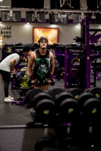 A person is engaged in weightlifting at a gym. They are wearing headphones and a tank top with a graphic design, standing among various gym equipment. Another individual is visible in the background, setting up weights. The environment includes purple and black gym machinery and weights, creating a focused workout atmosphere.