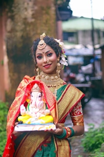 A woman dressed in traditional Indian attire with intricate jewelry holds a colorful statue of Lord Ganesha. She stands in an outdoor setting with a blurred background of greenery and buildings. Her attire includes a vibrant saree with red and gold patterns.