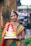 A woman dressed in traditional Indian attire with intricate jewelry holds a colorful statue of Lord Ganesha. She stands in an outdoor setting with a blurred background of greenery and buildings. Her attire includes a vibrant saree with red and gold patterns.