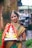 A woman dressed in traditional Indian attire with intricate jewelry holds a colorful statue of Lord Ganesha. She stands in an outdoor setting with a blurred background of greenery and buildings. Her attire includes a vibrant saree with red and gold patterns.