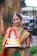 A woman dressed in traditional Indian attire with intricate jewelry holds a colorful statue of Lord Ganesha. She stands in an outdoor setting with a blurred background of greenery and buildings. Her attire includes a vibrant saree with red and gold patterns.