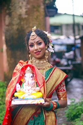 A woman dressed in traditional Indian attire with intricate jewelry holds a colorful statue of Lord Ganesha. She stands in an outdoor setting with a blurred background of greenery and buildings. Her attire includes a vibrant saree with red and gold patterns.